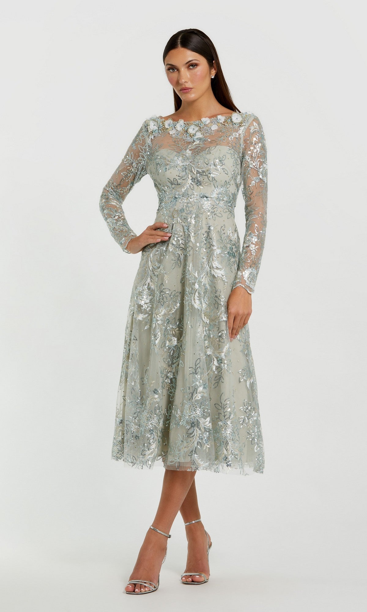 A woman wearing the Mac Duggal 68898 Midi Party Dress in Mist—a light blue, floral-embroidered dress featuring delicate lace and long sleeves—paired with silver strappy heels, standing against a plain white background.